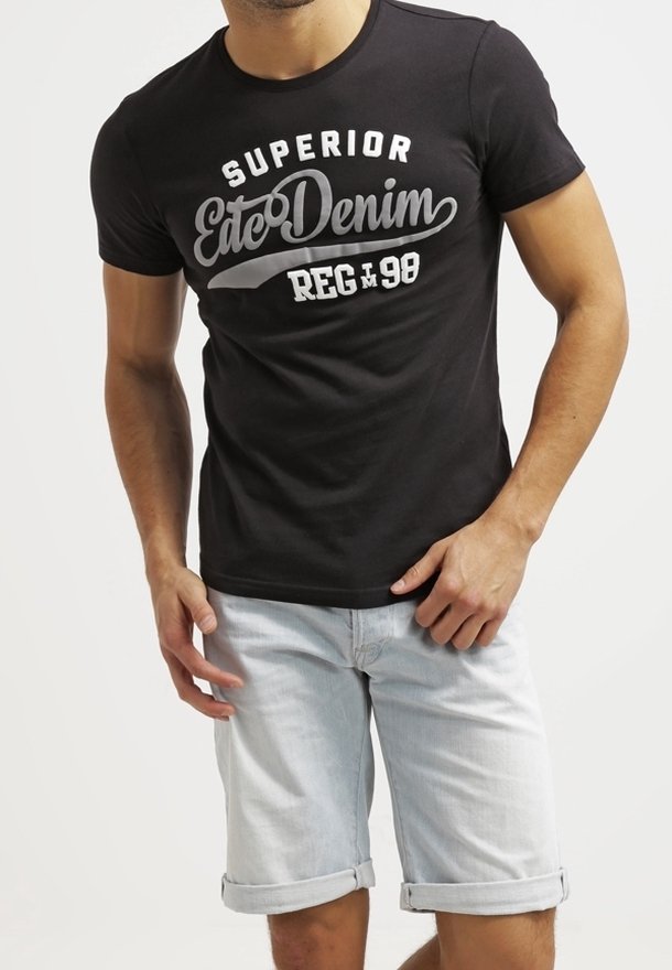 Homme portant un t-shirt noir avec texte blanc et un short en jean bleu clair retroussé sur un fond uni.