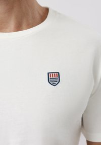 Κοντινό πλάνο ενός ατόμου που φοράει λευκό t-shirt με ένα μικρό έμβλημα ασπίδας με κόκκινες και μπλε ρίγες στο στήθος.