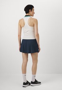 Top blanco sin mangas con cuello de contraste, combinado con una falda acampanada de color azul oscuro. La modelo lleva zapatillas deportivas negras y calcetines blancos.