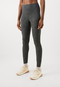 Leggings aus dunkelgrauem Stoff mit einem strukturierten Wirbelmuster, hoher Taille und einem figurbetonten Design. Kombiniert mit hellen Sportschuhen.