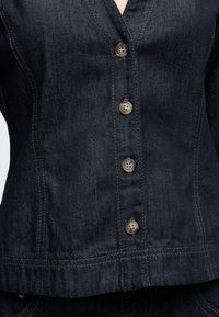 Σκούρο denim μπουφάν με πέντε ορατά κουμπιά, ραφές με αντίθεση και ραφές εφαρμογής στο μπροστινό μέρος του κορμού.