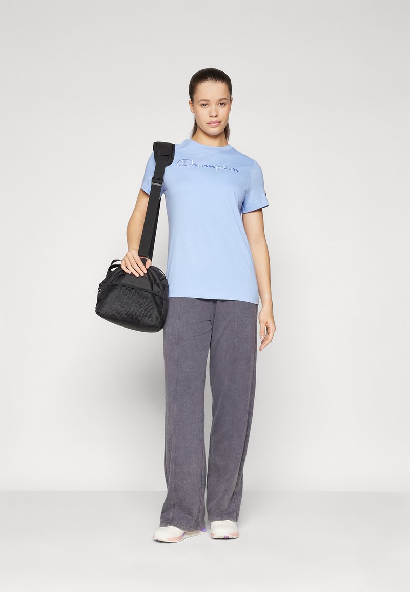 T-shirt azzurro con logo ricamato, abbinato a pantaloni grigi a gamba larga. Borsa nera con tracolla regolabile, dalla forma rotonda.