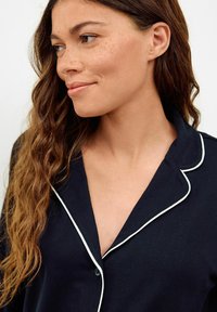 Woman with long wavy hair and freckles wearing a dark navy pajama top with white piping, looking to her left with a slight smile.