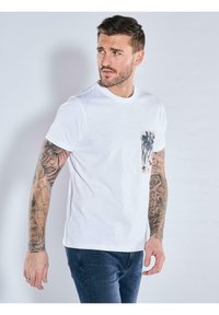 White cotton t-shirt with a round neckline and short sleeves. Features a textured pocket on the left with a dark, patterned accent.