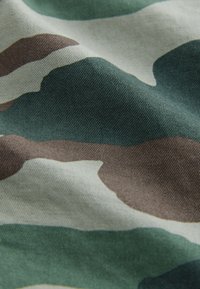 Tarnstoff mit einer Mischung aus Grün, Beige und Braun in unregelmäßigen, wellenförmigen Mustern und einer strukturierten, gewebten Oberfläche.