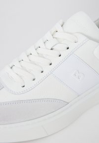 Sneaker bianca con tomaia in pelle liscia, dettagli in suede, punta tonda, lacci bianchi piatti e un piccolo logo in rilievo sul lato.