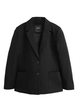 Ung pige med krøllet hår iført en sort blazer over en hvid skjorte med knapper, stående mod en ensfarvet lys baggrund.