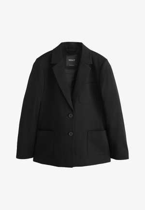 Ung pige med krøllet hår iført en sort blazer over en hvid skjorte med knapper, stående mod en ensfarvet lys baggrund.