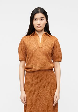 Naine, kes kannab sobivat roostepunast lühikeste varrukatega kootud pluusi kraega ja poolpikka seelikut, seisab ühevärvilise valge tausta ees.