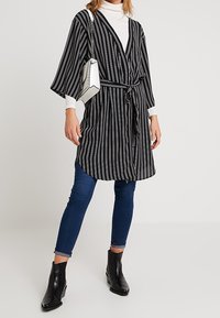 Gestreepte zwart-witte kimono-stijl wikkel met brede mouwen, vergezeld van een witte handtas en zwarte enkellaarsjes, gedragen over blauwe spijkerbroeken.