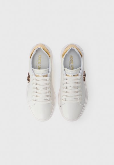Sneakers bianchi in pelle con accenti dorati sul tallone, lacci piatti e una caratteristica decorativa sul lato; texture liscia, design minimalista.