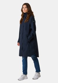 Cappotto lungo blu navy con colletto alto, zip frontale e tasche laterali; presenta una vestibilità comoda e una leggera curvatura all'orlo.