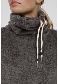 Grå, fluffig fleecepullov med hög krage, försedd med krämfärgade dragsnören och trädetaljer. Mjuk struktur och avslappnad design.