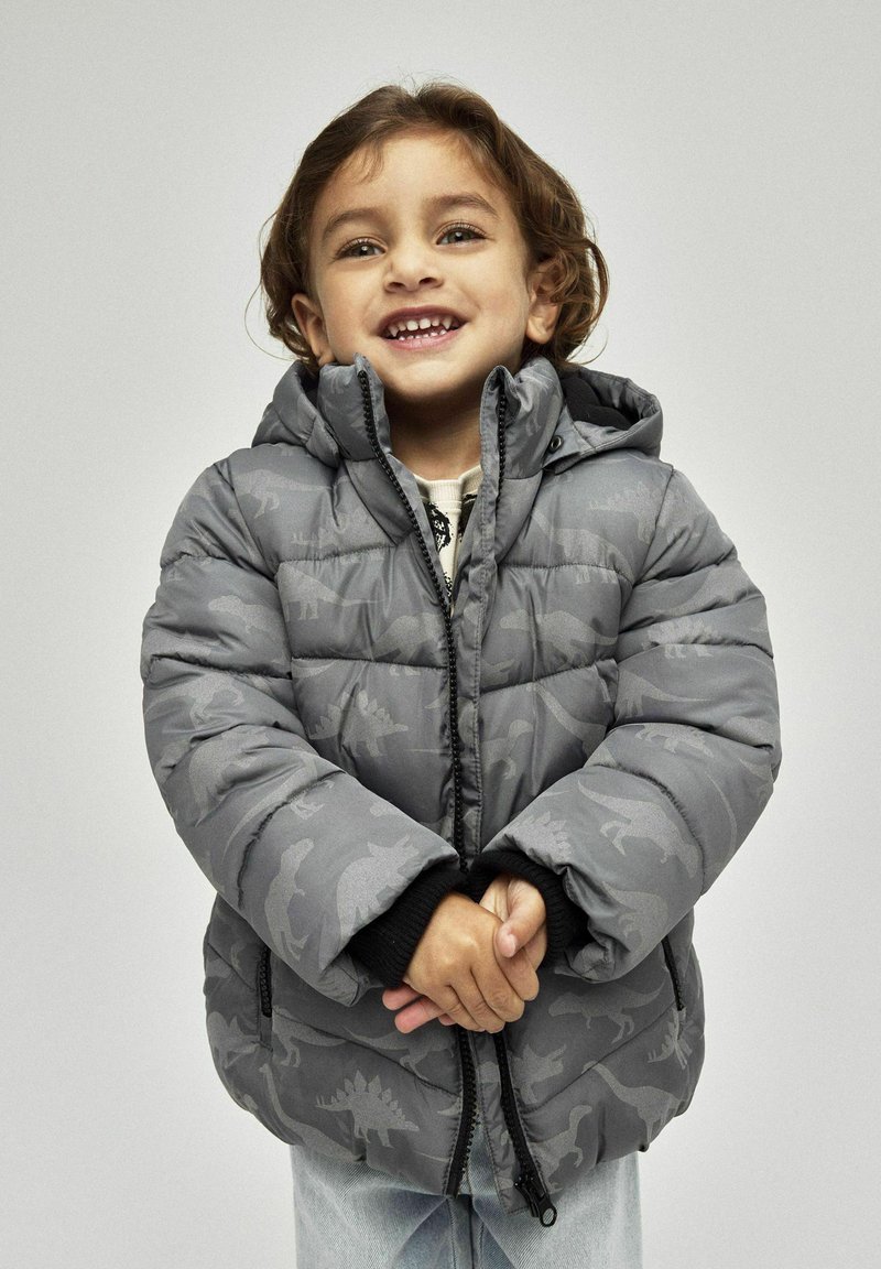 Graue Pufferjacke mit Dinosauriermustern, Kapuze und schwarzen Bündchen. Reißverschluss vorne und gestepptes Material, getragen von einem Kind.