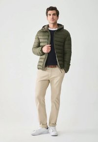 Man wearing green puffer jacket over navy sweater, beige pants with brown belt, and white sneakers, standing against a plain light background.