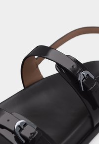 Κοντινό πλάνο ενός γυαλιστερού μαύρου σανδάλι με ρυθμιζόμενους ιμάντες και ασημένιες αγκράφες σε μια λεία μαύρη σόλα.