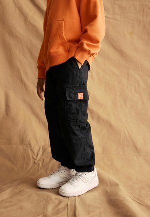 Oranžs kapučīnu džemperis ar priekšējo kabatu, melni cargo bikses ar sānu kabatām un baltas sneakers ar logotipa detaļām, uz beige fona.