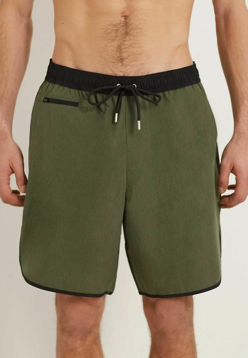 Pantaloni da bagno verdi con vita elastica nera, cordoncino regolabile e tasca laterale bordata di nero. Tessuto leggero e strutturato.
