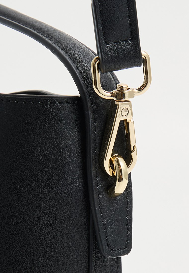 Détail d'un sac à main en cuir noir avec un fermoir et du matériel en métal doré, des coutures visibles le long des bords et une texture lisse.