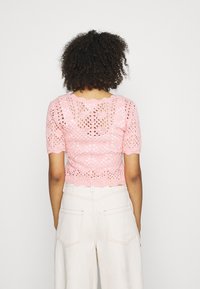 Rosa gestricktes Oberteil mit kurzen Ärmeln, das ein offenporiges Design und einen gewellten Saum aufweist. Die Rückansicht zeigt ein spitzenähnliches Muster und Struktur.