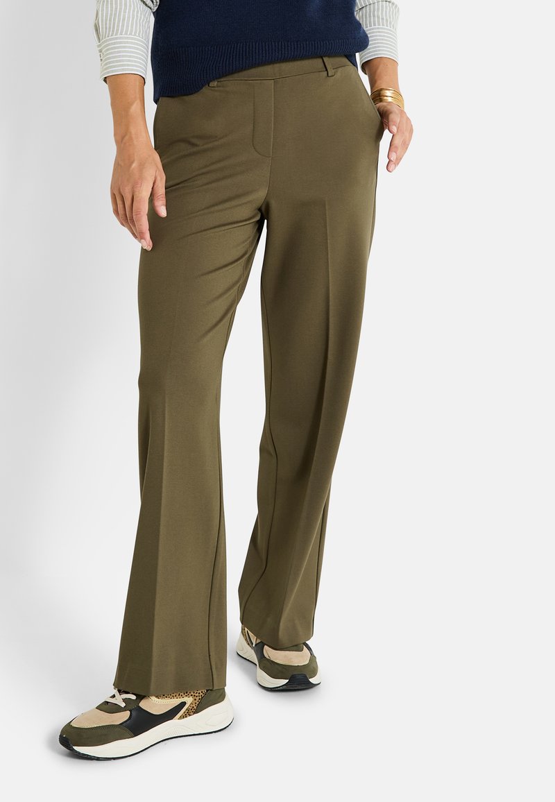 Pantalons larges vert olive en tissu lisse, dotés de poches latérales et d'un pli subtil à l'avant, associés à des baskets beiges et noires.