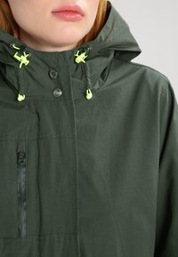 Grön vattentät jacka med huva, med en ficka med dragkedja, tryckknappar och ljusgröna dragsnoddar. Slätt, lättviktigt tyg.