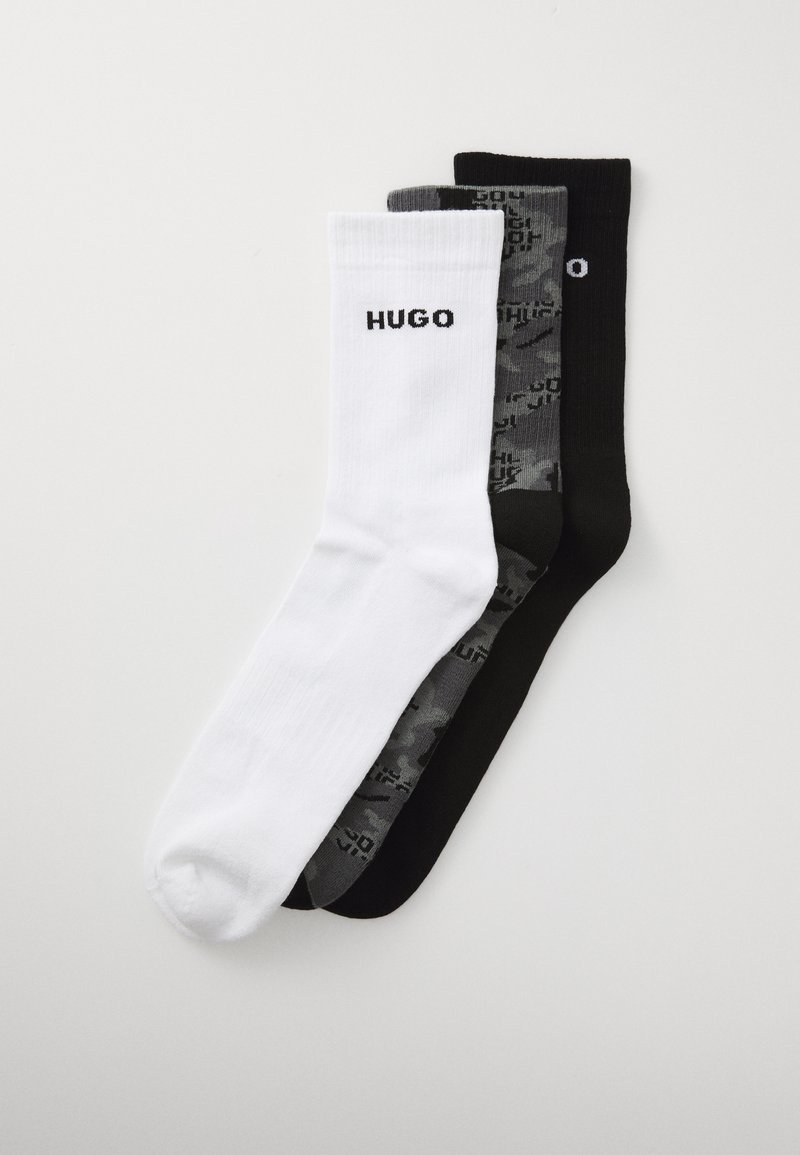 Trois paires de chaussettes de sport en blanc, camouflage gris et noir, chacune avec le texte "HUGO" sur le poignet, disposées à plat sur un fond blanc.