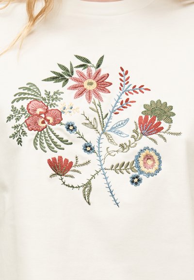 Κεντήματα με λουλούδια σε ροζ, κόκκινους, μπλε και πράσινους τόνους πάνω σε κρεμ ύφασμα.