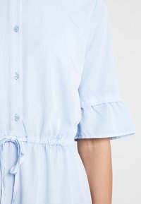Jasnoniebieska sukienka z krótkim rękawem, z zapięciem na guziki, falbaniastymi rękawami i elastycznym paskiem w talii do regulacji. Miękka tekstura o gładkim wykończeniu.