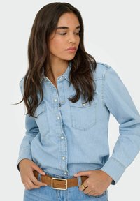 Jasnoniebieska koszula dżinsowa z ozdobnymi guzikami, spiczastym kołnierzykiem, dwoma kieszeniami na piersi i długimi rękawami, noszona z brązowym paskiem i niebieskimi dżinsami.