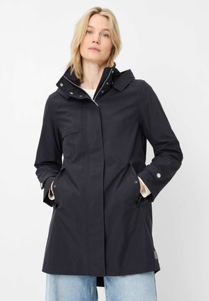 Frau trägt eine schwarze, knielange Regenjacke mit Fronttaschen und hohem Kragen, kombiniert mit hellblauen Jeans, stehend vor weißem Hintergrund.