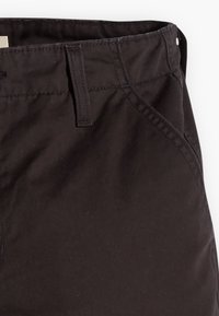 Gros plan de la partie avant supérieure d’un pantalon brun foncé montrant la ceinture, les passants de ceinture et la couture diagonale de la poche avant.