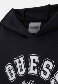 Sweat à capuche noir avec "GUESS" en grandes lettres blanches ornées d'accents en cristaux. Texture douce et design sportif.