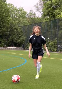 Juoda sportinė marškinė ir šortai, balti dryžiai, iki kelių balti kojinių, neoniniai geltoni sportbačiai ir raudonai balta futbolo kamuolys ant žolės aikštės.