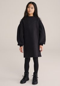 Juoda didelio dydžio džemperio suknelė su nukritusiomis pečių linijomis, ribinėmis rankogalių apyrankėmis ir apvaliu iškirptu kaklu, derinama su juodomis tampriomis kelnėmis ir batais su raišteliais iki kulkšnies.