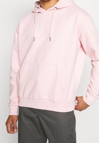 Różowa bawełniana bluza z kapturem z przednią kieszenią, ściągacze przy mankietach, i wyszywane logo na biało. Sznurki z metalowymi końcówkami. Miękka tekstura.