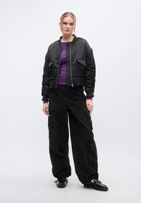 Μαύρο μπουφάν τύπου bomber με υφή καπιτονέ, δύο μπροστινές τσέπες. Φοριέται πάνω από μωβ μπλούζα με μακριά μανίκια και μαύρο παντελόνι cargo, με φερμουάρ και άνετη εφαρμογή.