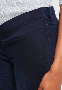 Mörkblå jeans med en elastisk midja, som har en slät yta och en klassisk femfickdesign. Närbild visar detaljerna vid midjan.