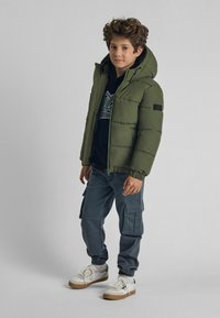 Giacca a vento verde con cappuccio, zip frontale e polsini elastici sopra una felpa nera. Abbinata a pantaloni cargo grigi e sneakers bianche.