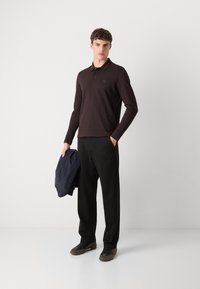 Brun langærmet polo med et lille logo, sorte bukser, holder en marineblå jakke og har mørke sneakers på en lys baggrund.