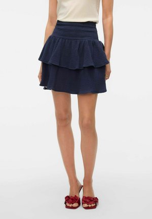 Mini-jupe à volants superposés de couleur bleu marine, fabriquée en tissu texturé. Taille élastique pour un meilleur confort et une plus grande flexibilité. Assortie à des chaussures ouvertes de couleur rouge.