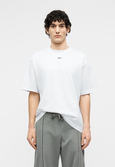 Νεαρός άνδρας με σγουρά μαλλιά φορώντας υπερμεγέθη λευκή μπλούζα με το κείμενο «OFF» και γκρι παντελόνι με κορδόνι, σε απλό ανοιχτό φόντο.