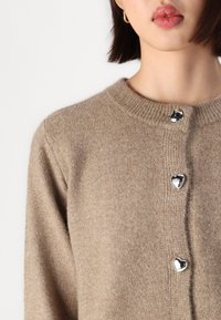 Cardigan de malha bege com decote redondo, apresentando três botões prateados em forma de coração e punhos canelados. Textura suave e corte descontraído.