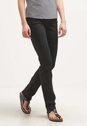 Osoba ubrana w czarne dopasowane spodnie i czarne sandały, stojąca ze skrzyżowanymi nogami na jednolitym białym tle.