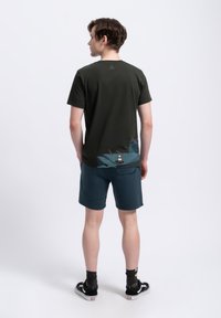 Maglietta a maniche corte verde scuro con un design grafico testurizzato vicino all'orlo, abbinata a pantaloni corti verde teal scuro e sneakers nere.