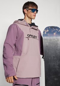 Paarse technische snowboardjas met capuchon, voorzien van zwart logo, ritsen en textuurpanelen. Vergezeld van een snowboard.