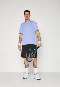 Ljusblå t-shirt med korta ärmar, svarta shorts, vita handledsband, håller en tennisklubba. Vita sneakers med blå accenter, synliga tatueringar.
