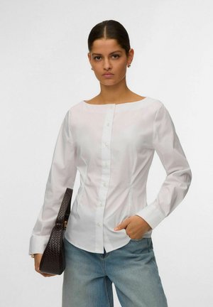 Naine kannab valget, keha jälgivast kangast nööbitavat pluusi ja helehalli teksaseid, hoides tumeda tekstuuriga käekotti ning ühe käega on taskus.