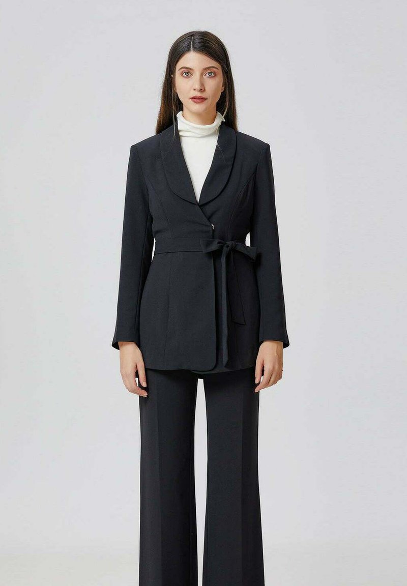 Femme avec de longs cheveux noirs portant un blazer noir ceinturé sur un turtleneck blanc et un pantalon large noir, se tenant contre un fond clair.
