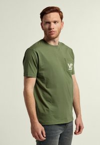 Camiseta de algodón verde con cuello redondo, mangas cortas y un bolsillo en el pecho que presenta un logo blanco. Textura suave. Corte estándar.